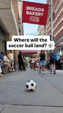 Red Breads Bakery sign hangs above a sidewalk with people walking and a soccer ball on the pavement