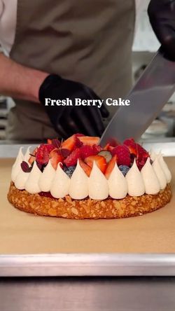 Fresh berry cake topped with cream and assorted berries being sliced in a bakery kitchen.