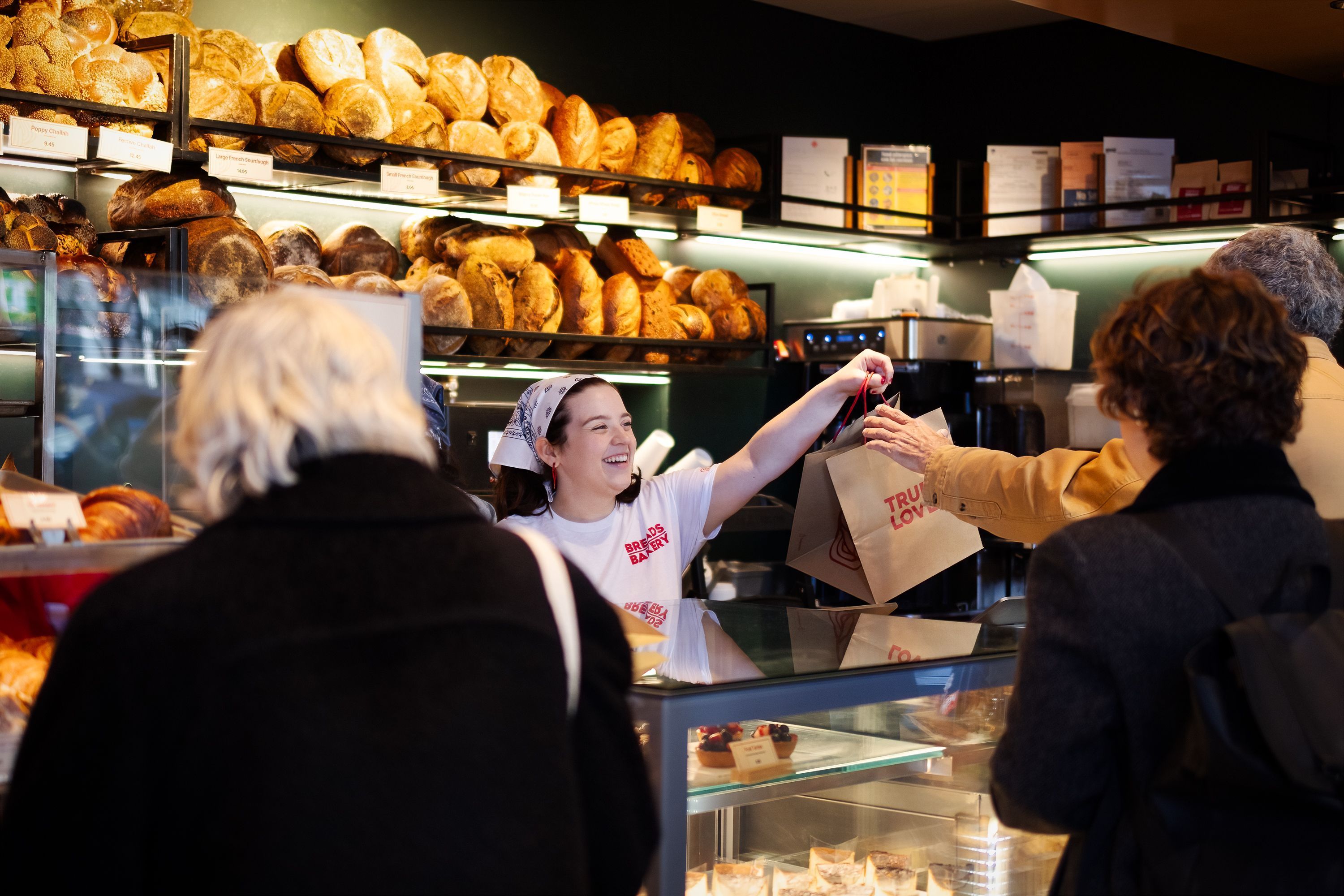 A bakery counter with various breads on display and customers being served takeaway bags.