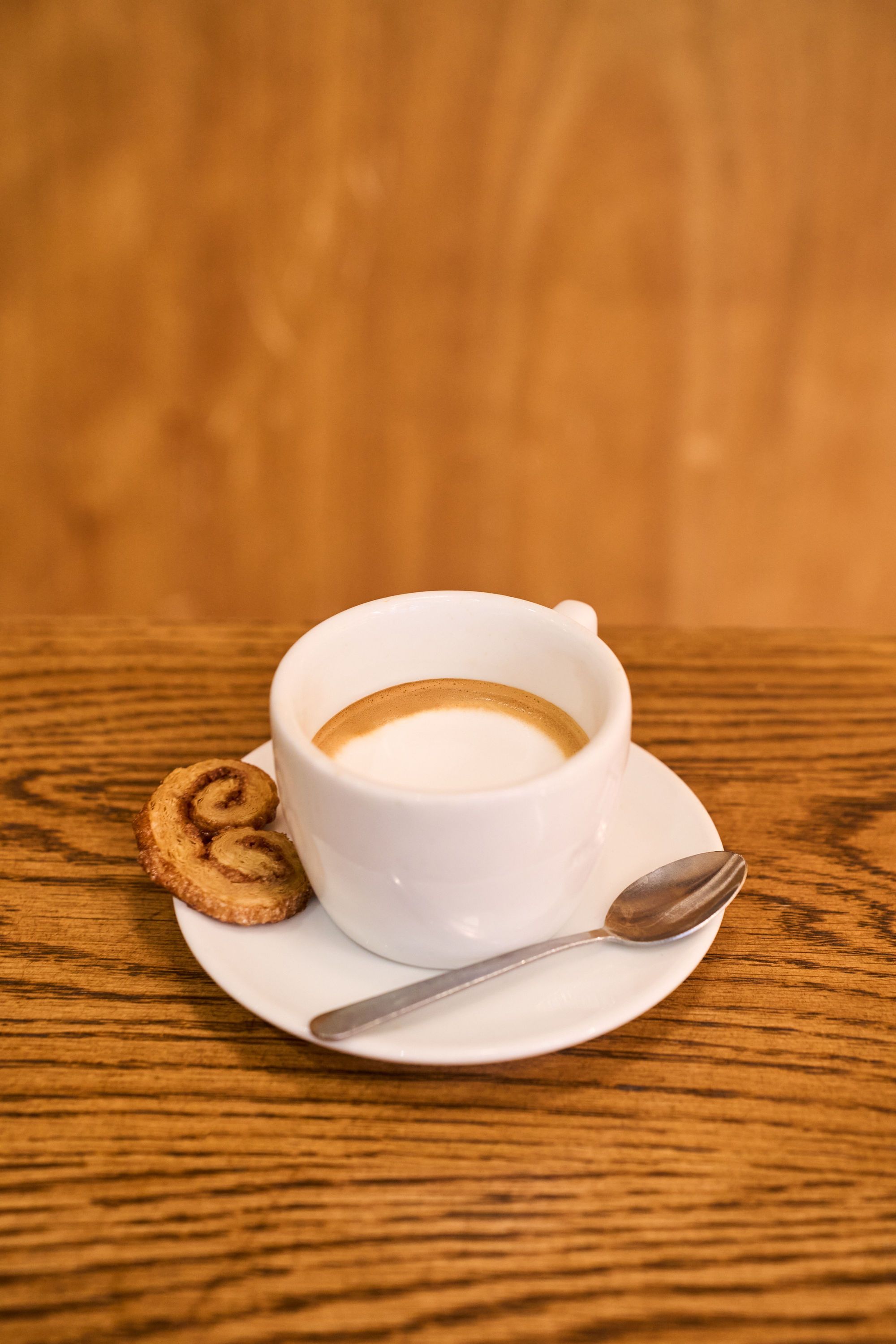 A cup of coffee on a white saucer accompanied by a pastry, set on a wooden table.