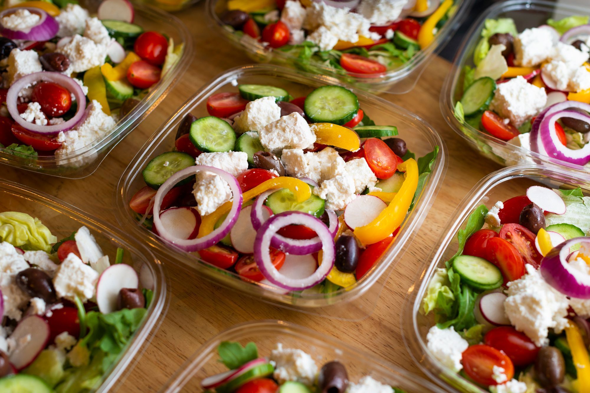 Fresh vegetable salads with tomatoes, cucumbers, olives, onions, and cheese in clear containers on a wooden surface